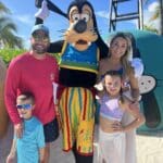 Family smiling with Goofy on Disney’s Castaway Cay private island, enjoying a sunny beach day in the Bahamas with palm trees and bright blue skies in the background.