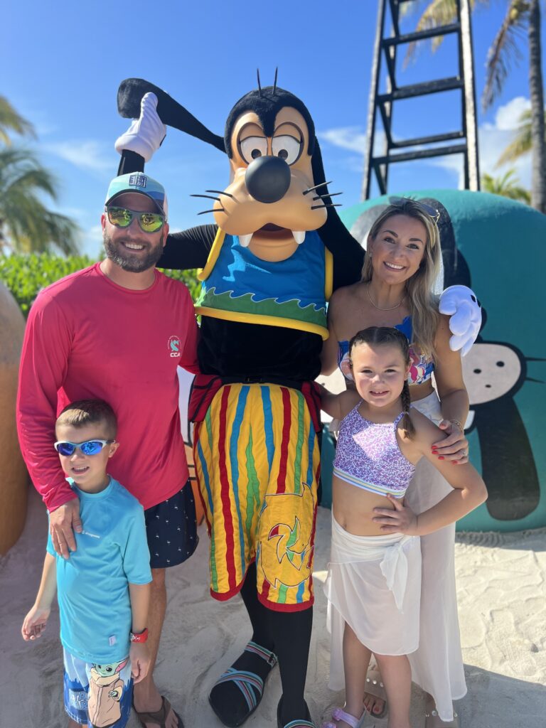Family smiling with Goofy on Disney’s Castaway Cay private island, enjoying a sunny beach day in the Bahamas with palm trees and bright blue skies in the background.