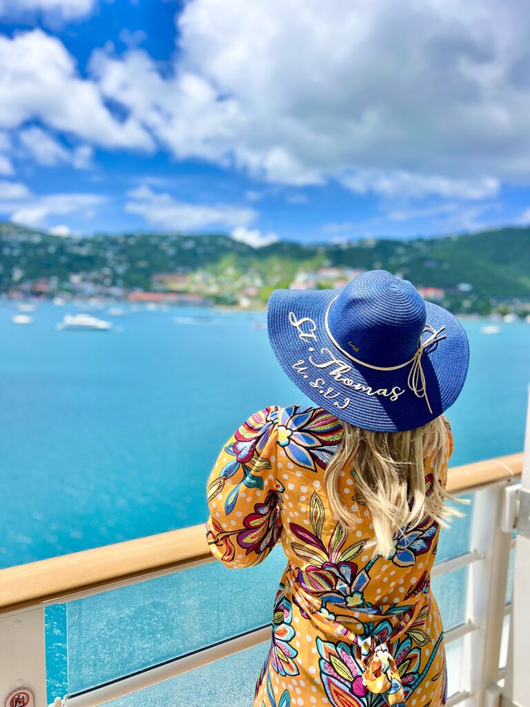 Woman in a floral dress and blue sun hat looking out from a Disney Cruise Line balcony at the turquoise waters and lush hills of St. Thomas in the Eastern Caribbean.