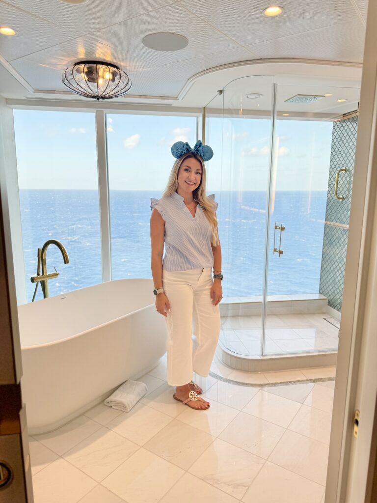 Smiling woman wearing Disney ears stands in a stunning luxury bathroom aboard the Disney Treasure cruise ship, featuring a soaking tub and glass shower with panoramic ocean views.