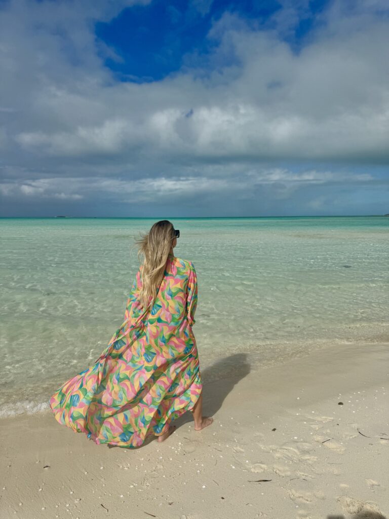 Woman in a colorful flowy dress walking along the turquoise shoreline of Grace Bay Beach in Turks and Caicos under a bright blue sky.
