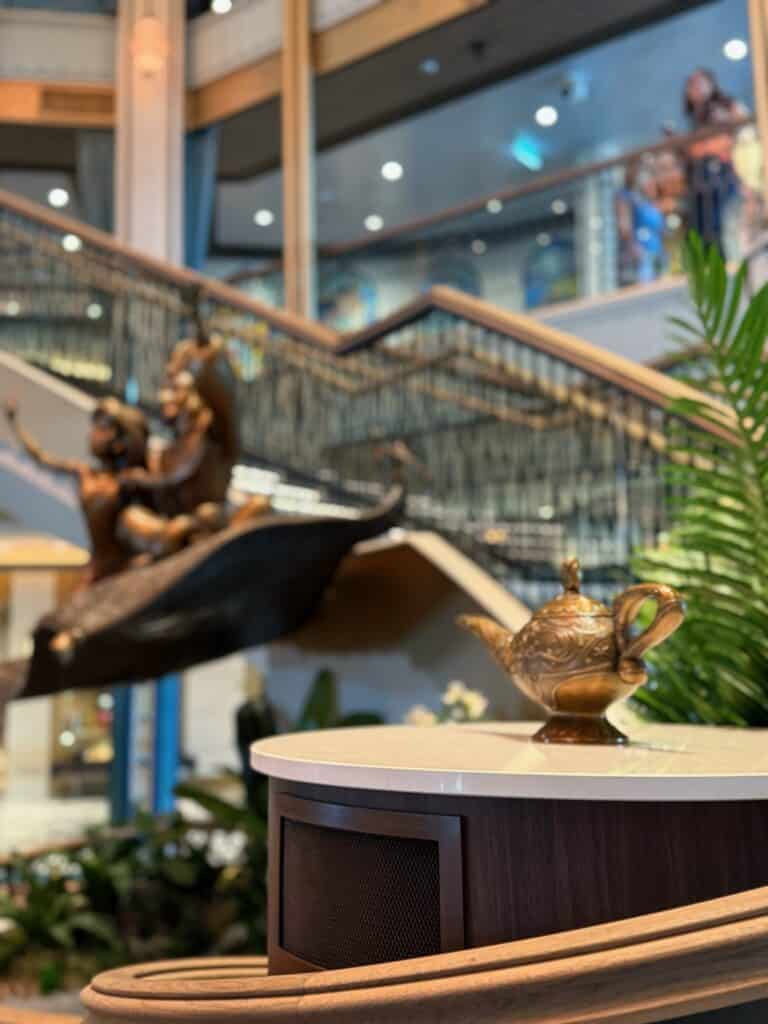 The grand atrium of the Disney Treasure cruise ship featuring the Aladdin and Jasmine statue with a golden magic lamp in the foreground.