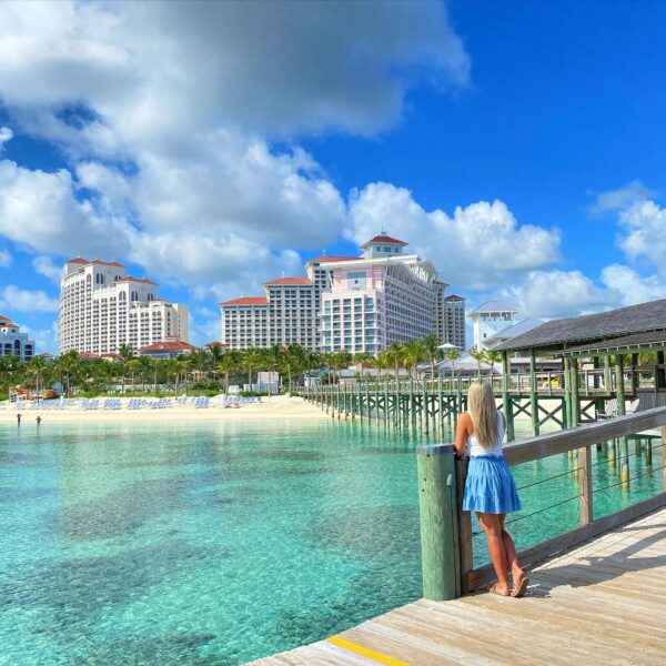 Woman in a blue skirt standing on a boardwalk overlooking the clear turquoise water and iconic towers of Baha Mar Resort on Cable Beach in Nassau, Bahamas.