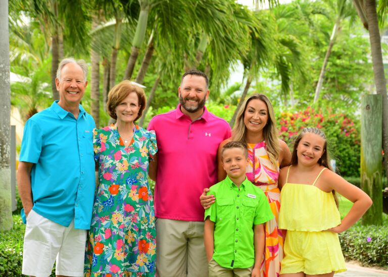 Smiling family of six posing together among lush palms and tropical flowers at Beaches Turks & Caicos, dressed in bright colorful outfits during a Caribbean family vacation.