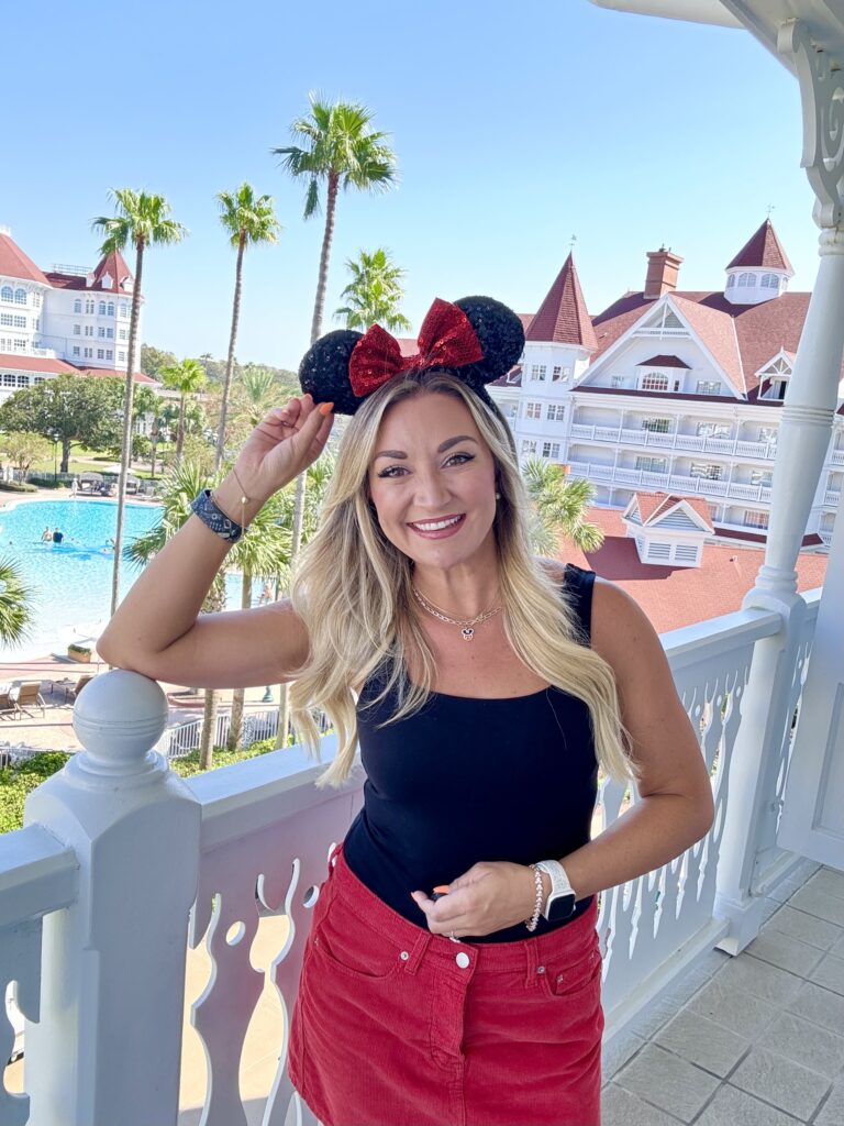 Woman smiling in Minnie ears on the balcony at Disney’s Grand Floridian Resort, with palm trees, a pool, and the iconic red-roofed Victorian buildings in the background.