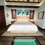 A luxurious overwater bungalow bedroom at Sandals South Coast in Jamaica, featuring a beautiful glass floor panel showcasing the turquoise water below, a plush king bed, and elegant tropical decor.