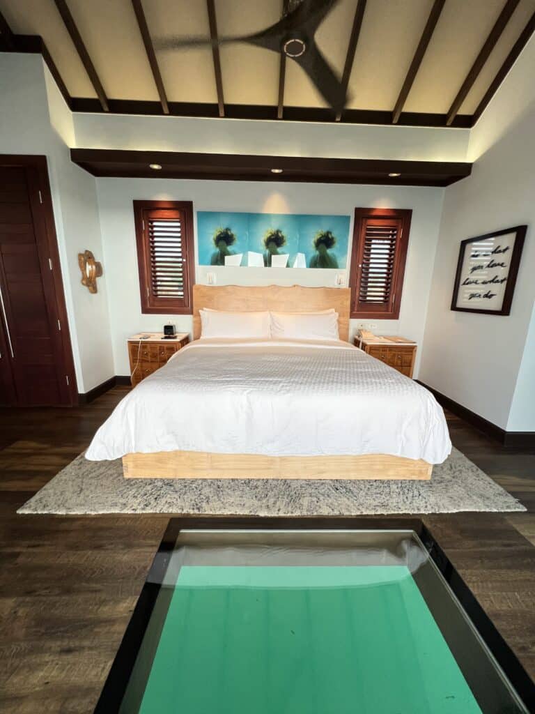 A luxurious overwater bungalow bedroom at Sandals South Coast in Jamaica, featuring a beautiful glass floor panel showcasing the turquoise water below, a plush king bed, and elegant tropical decor.
