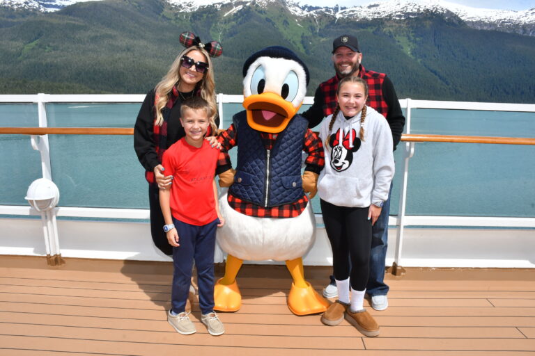 Family posing with Donald Duck in his Alaskan plaid outfit on the top deck of the Disney Wonder, with stunning snow-capped mountains and turquoise water in the background during a Disney Alaska Cruise.