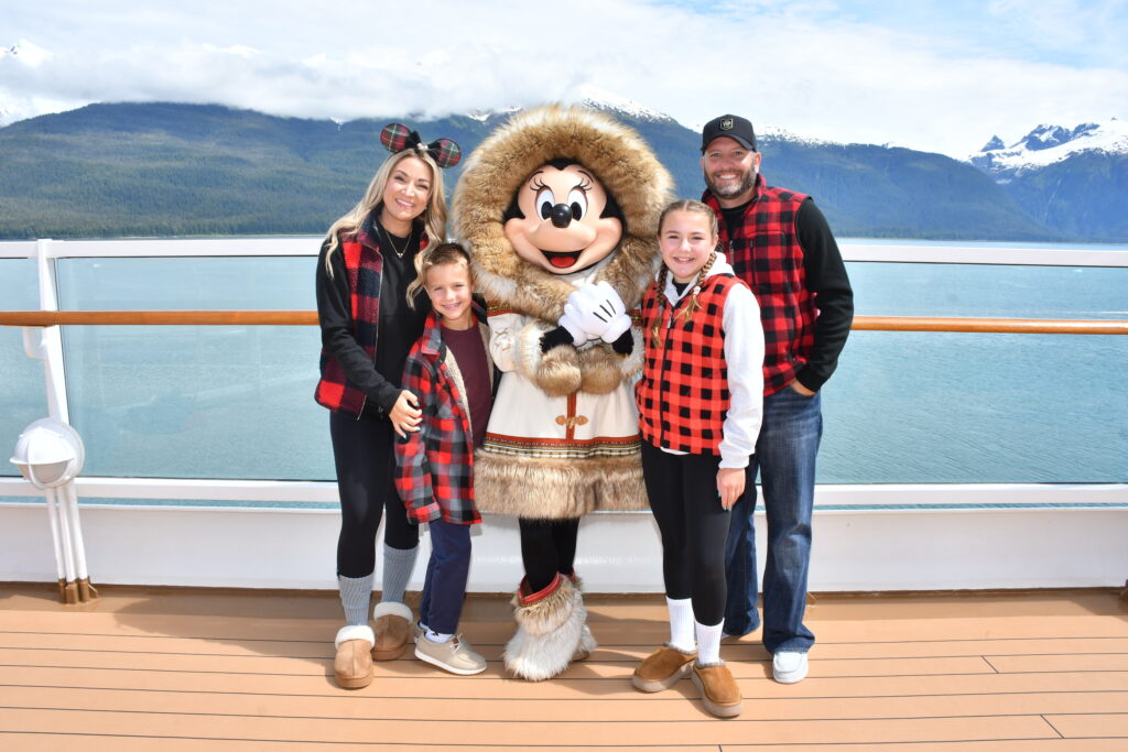 Family posing with Minnie Mouse in her cozy fur-trimmed Alaskan outfit on the top deck of the Disney Wonder, with turquoise waters and snow-capped mountains in the background during a Disney Alaska Cruise.