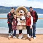 Family posing with Minnie Mouse in her cozy fur-trimmed Alaskan outfit on the top deck of the Disney Wonder, with turquoise waters and snow-capped mountains in the background during a Disney Alaska Cruise.