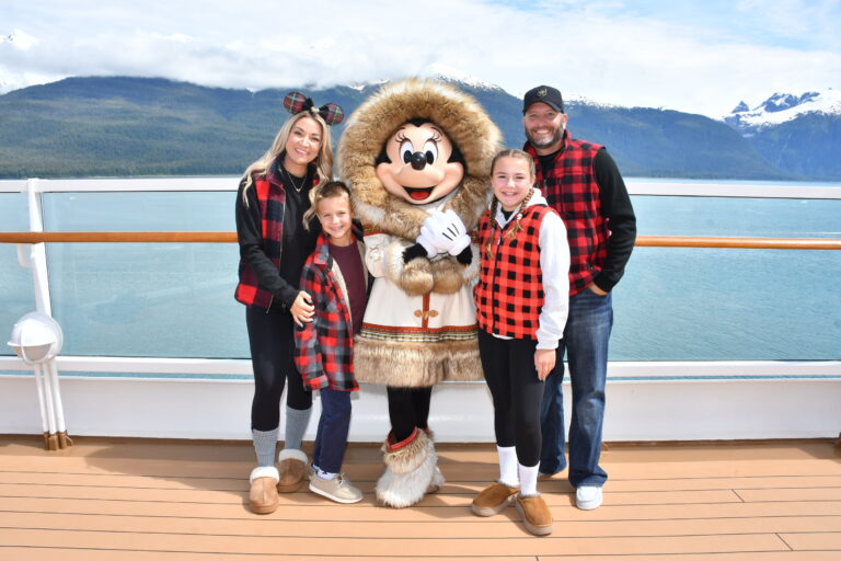 Family posing with Minnie Mouse in her cozy fur-trimmed Alaskan outfit on the top deck of the Disney Wonder, with turquoise waters and snow-capped mountains in the background during a Disney Alaska Cruise.