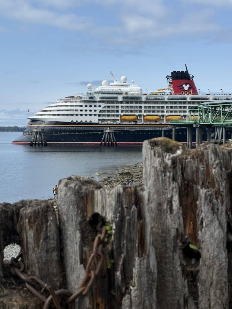 The Disney Wonder cruise ship docked at Icy Strait Point, Alaska, during a Disney Alaska Cruise, framed by rustic wooden pilings along the shoreline.