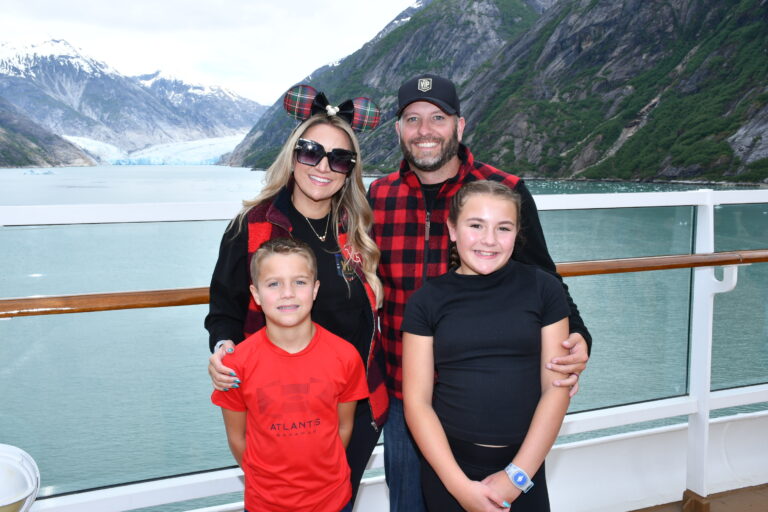 Family of four posing on the deck of the Disney Wonder with a stunning glacier and snow-capped mountains in the background during an Alaska cruise.
