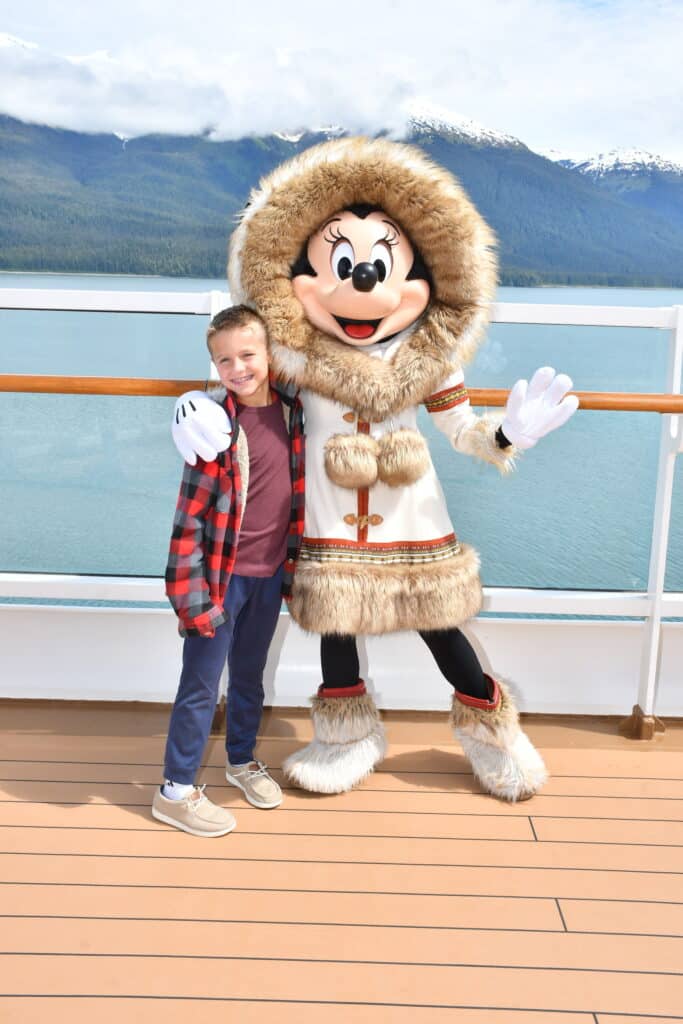 Young boy posing with Minnie Mouse in a fur-trimmed white parka aboard the Disney Wonder during an Alaska cruise, with snowy mountains in the background.