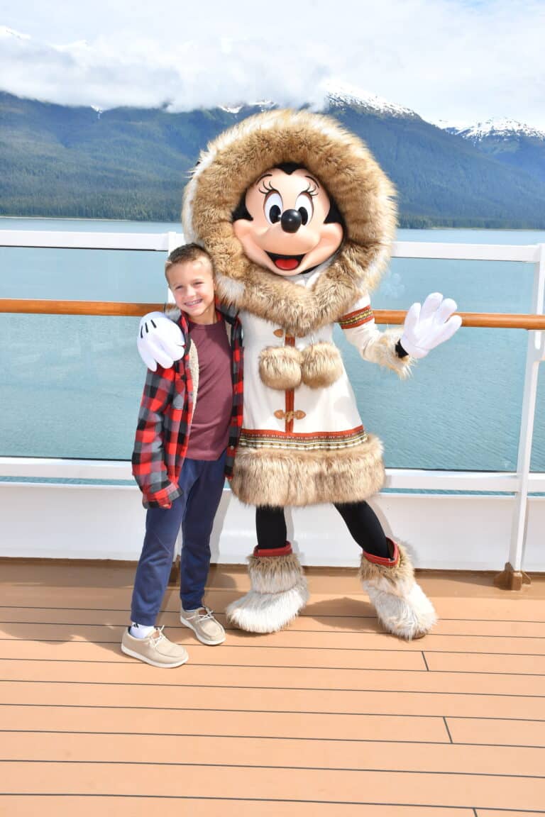 Young boy posing with Minnie Mouse in a fur-trimmed white parka aboard the Disney Wonder during an Alaska cruise, with snowy mountains in the background.