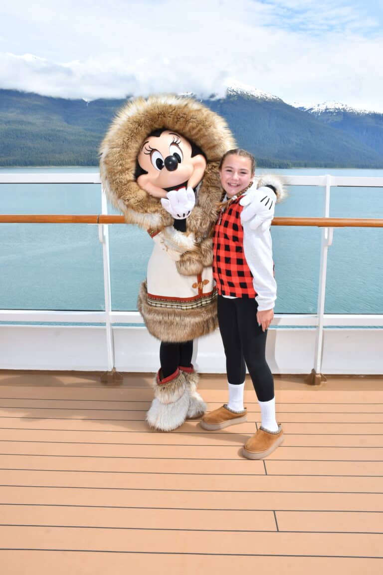 Paisley smiles alongside Minnie Mouse dressed in a fur-lined parka on the deck of the Disney Wonder with Alaska’s snow-capped mountains and water in the background.