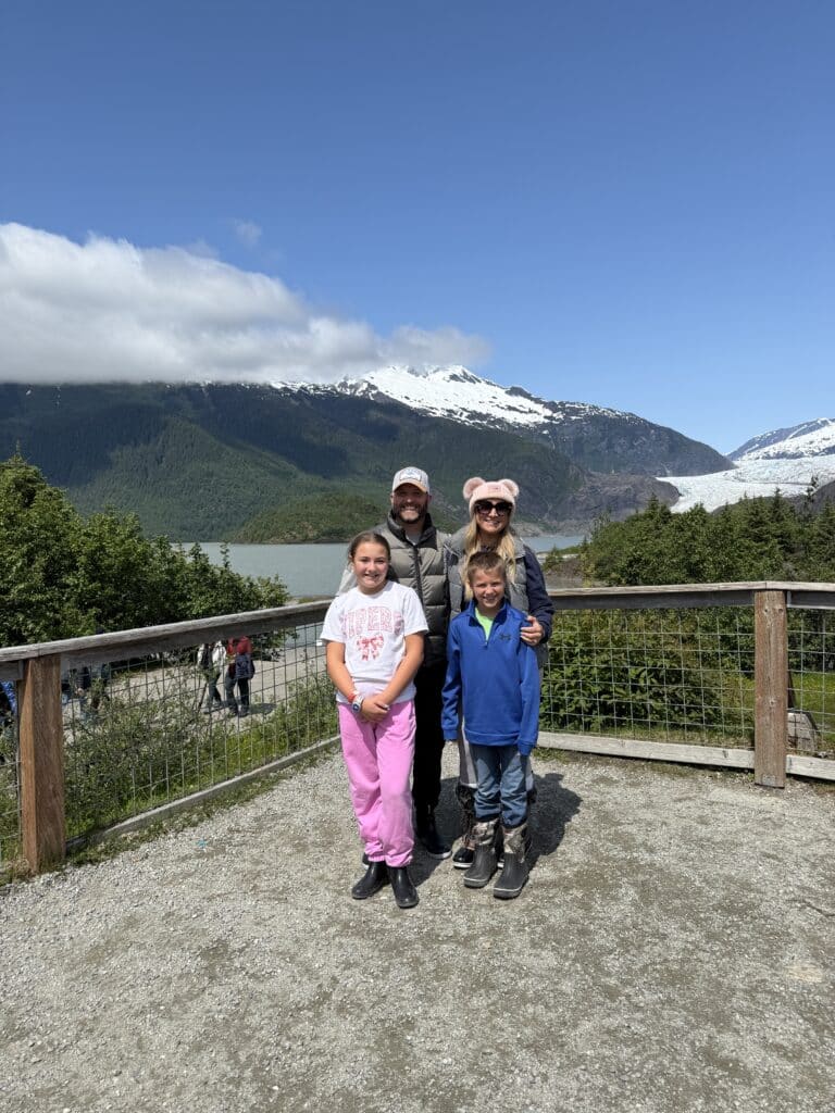 Family posing in front of the stunning Mendenhall Glacier in Juneau, Alaska, with snow-capped mountains, lush greenery, and a bright blue sky in the background.