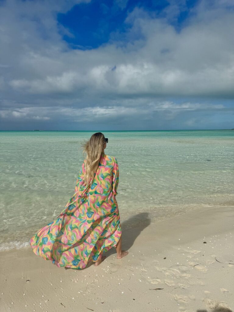 Woman in a colorful flowy dress walking along the turquoise shoreline of Grace Bay Beach in Turks and Caicos under a bright blue sky.