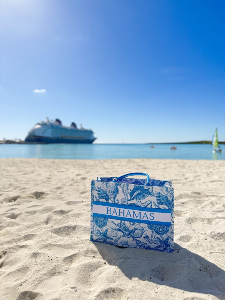 Castaway Cay beach bag on the sand with the Disney Fantasy cruise ship anchored in the turquoise waters of Disney’s private island.